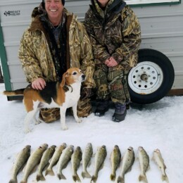 Two people in winter clothing kneeling beside a dog and a row of nine fish on ice, with a building and a tire in the background.