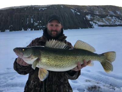 A person dressed in winter clothing holds a large fish standing on a snowy, frozen water body with rocky cliffs in the background.