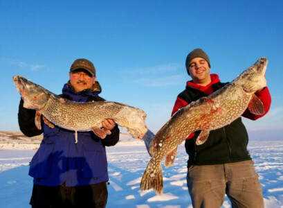 Two people standing on a snowy landscape, each holding a large fish. Both are dressed warmly and appear pleased with their catch.