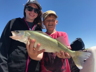 Two people standing with a large fish one of them is holding, both smiling under a clear blue sky.