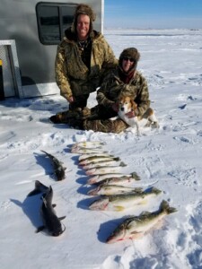 Two people in winter clothing kneeling on snow with a dog and several fish lined up in front of them, next to a trailer.