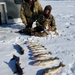 Two people in winter clothing kneeling on snow with a dog and several fish lined up in front of them, next to a trailer.