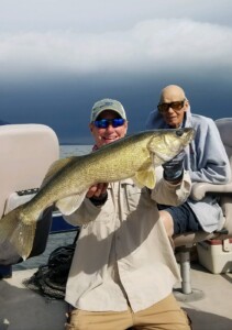 Two men on a boat; one man kneels holding a large fish, while the other sits in a chair, both wearing sunglasses.
