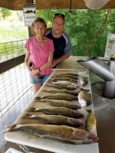Two people stand behind a table displaying a row of freshly caught fish, with a cleaning station in the background.