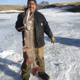 A man stands on ice, holding a large fish with both hands. He is wearing winter clothing, including a hat, gloves, and boots.