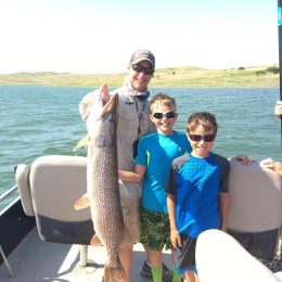 Three people on a boat, with one holding a large fish and two children beside them. The water and shoreline are visible in the background.