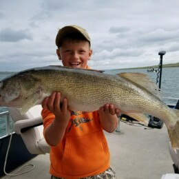 A young person wearing an orange hoodie proudly holds a large fish on a boat with water and another boat visible in the background.