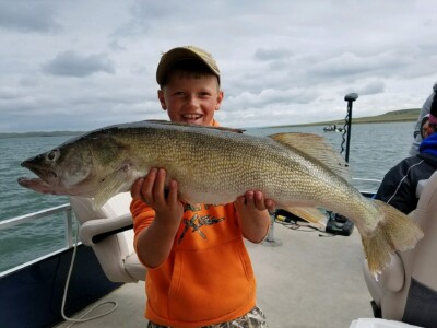 A young person wearing an orange hoodie proudly holds a large fish on a boat with water and another boat visible in the background.