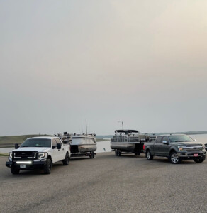 Two trucks towing boats parked on a lakeshore, preparing for a day of adventure. One boat appears to be set up for walleye fishing, while the other is a leisurely pontoon boat.