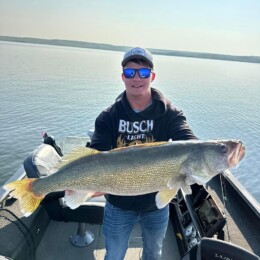 A person wearing a cap and sunglasses proudly displays a large fish, likely caught during a thrilling day of walleye fishing, on a boat with a calm body of water in the background.