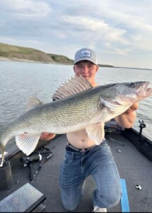 Person kneeling on a boat and holding a large walleye with scenic water and hills in the background, capturing an unforgettable walleye fishing experience.