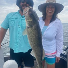 Two people wearing sun hats, smiling on a boat, holding a large walleye they caught during an exhilarating fishing trip, with a cloudy sky in the background.