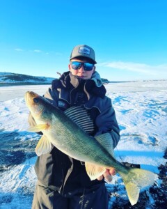A person proudly displaying a large fish caught during a thrilling walleye fishing expedition, standing on a snow-covered surface with a clear blue sky in the background.