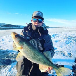 A person proudly displaying a large fish caught during a thrilling walleye fishing expedition, standing on a snow-covered surface with a clear blue sky in the background.
