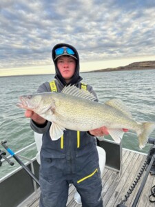 Person wearing a hooded jacket and holding a large walleye on a boat, with water and cloudy sky in the background.