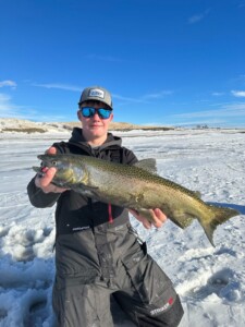 Person in winter clothing holding a large fish while standing on a snowy landscape under a clear blue sky, enjoying a successful day of Walleye fishing.