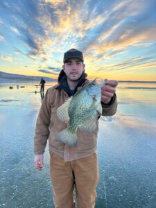 Person in a brown jacket and cap holding a walleye on an icy lake with sunset sky in the background.