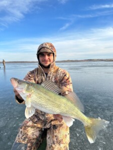 A person in camouflage gear proudly holds up a large fish caught while walleye fishing on a frozen lake, with another person in the background.