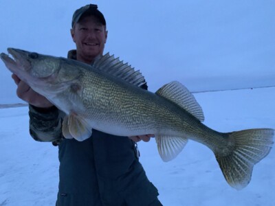 Person in outdoor gear holding a large walleye against a snowy backdrop with a slight smile.