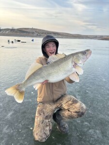 Person kneeling on ice holds a large walleye with a look of excitement, wearing a hooded jacket and camouflage pants.