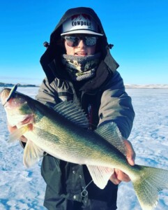 Person wearing outdoor gear holds up a large walleye on a snowy, frozen lake. They are dressed warmly with a hat, sunglasses, and jacket, embodying the thrill of walleye fishing in winter conditions.