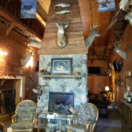 A rustic room featuring a stone fireplace, hunting trophies, mounted deer heads, wooden walls, and two cushioned chairs in front.