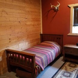 A bedroom with a single wooden bed, multicolored striped bedding, a wooden nightstand, a window, and a deer antler mounted on the wall.