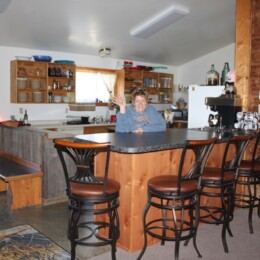 A person waves from behind a kitchen counter in a rustic room with bar stools, wooden cabinets, and aviation-themed decor.