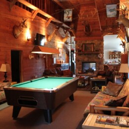 A rustic living room with a pool table, sofas, a stone fireplace, wood walls, and mounted deer trophies adorning the walls.