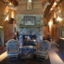 Rustic room with a stone fireplace, mounted deer heads, two armchairs, a rifle cabinet, and a bar area.