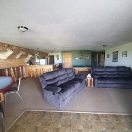 A living room with grey recliner sofas, a round card table, deer head mounts on the wall, and a small bar counter in the background.