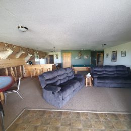 A living room with grey recliner sofas, a round card table, deer head mounts on the wall, and a small bar counter in the background.