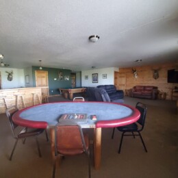 A large room features a poker table, pool table, couches, and mounted deer heads on the walls. The space has wooden paneling and muted colors.