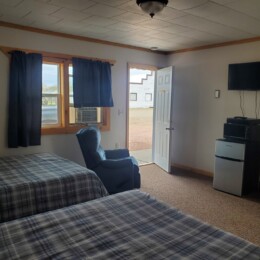 A room with two plaid-covered beds, a blue armchair, TV, mini fridge, and an open door revealing an outdoor view.