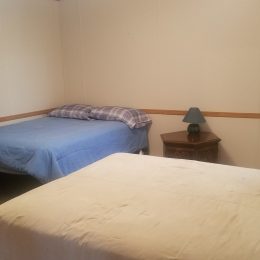 A bedroom with two beds, a nightstand with a lamp, blue curtains, and light-colored walls. The beds have blue and beige bedding.