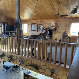 A wooden cabin interior with mounted animal heads and pelts on walls, a deer statue, and a railing overlooking a lower level dining area.