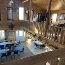 A spacious cabin interior with wooden walls, mounted animal heads, a dining area with tables and chairs, and an upper loft.