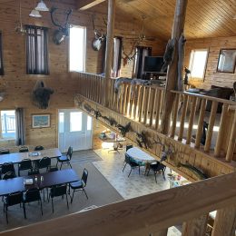 A spacious cabin interior with wooden walls, mounted animal heads, a dining area with tables and chairs, and an upper loft.