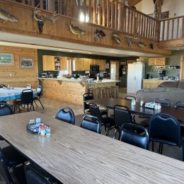 Spacious lodge interior with wooden decor, dining tables, a kitchen area, and a living space. Taxidermy on walls and rustic furniture.