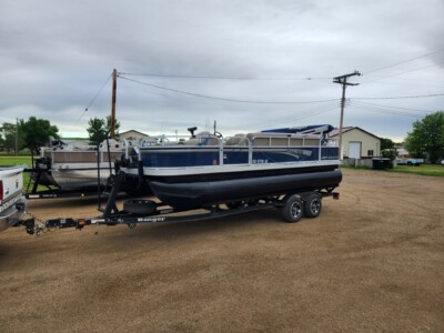 A blue and white pontoon boat on a trailer is parked outdoors on a dirt lot with other boats and buildings in the background.